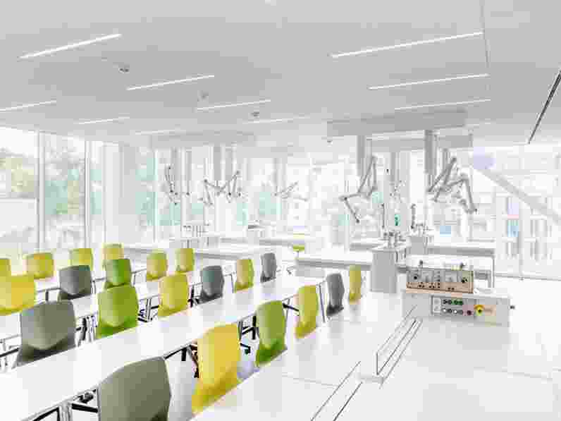 Moderní třída s velkými okny, jasným přirozeným světlem a barevnými židlemi uspořádanými v řadách. Strop obsahuje pokročilé osvětlení a bílé zavěšené akustické panely s laboratorním vybavením a odsávacími rameny namontovanými na stropě.