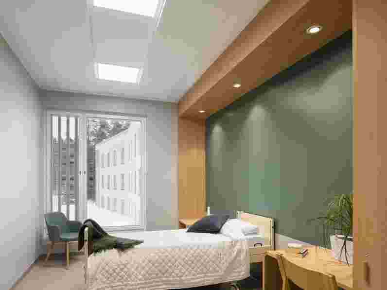Bílý akustický celoplošný podhled  v pokoji pro pacienty psychiatrické kliniky s dřevěnými detaily interiéru a nábytkem a tmavě zelenou stěnou