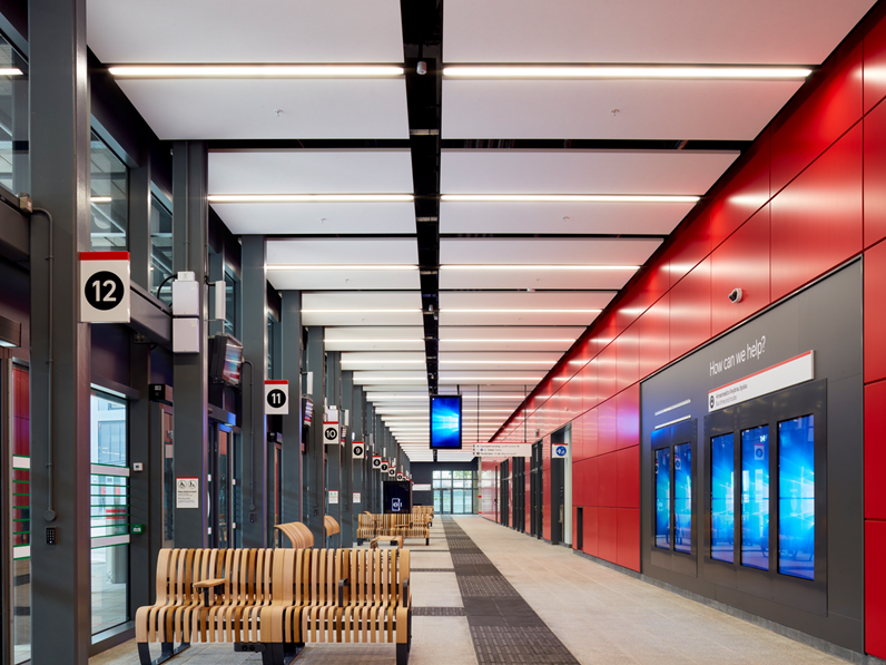 Modern bus station interior with high ceilings, red and gray walls, digital information screens, and wooden benches. Bright lighting illuminates the space, with a long corridor visible in the background. Signage with numbers hangs from the ceiling.