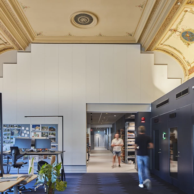 A modern office space features high decorative ceilings with ornate designs. There are workstations with computers and shelves filled with books and magazines. Two people walk in the background near a hallway.